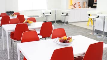 A conference room with white tables and red chairs, a projector screen, and a kitchenette in the background.