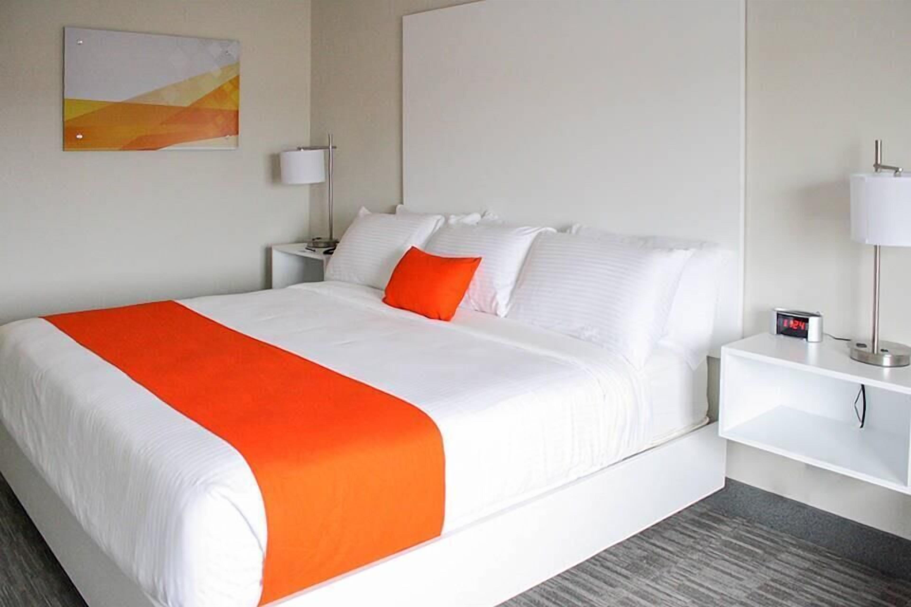A hotel room with a large bed, white bedding, an orange throw blanket, a nightstand with a lamp, and a painting on the wall.