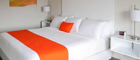 A hotel room with a large bed, white bedding, an orange throw blanket, a nightstand with a lamp, and a painting on the wall.