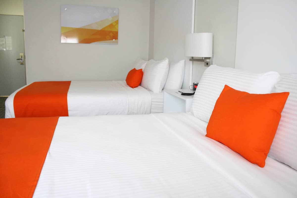 A hotel room with two beds, each with white bedding and orange pillows. There is a nightstand with a lamp and a small table. A painting is visible on the wall.
