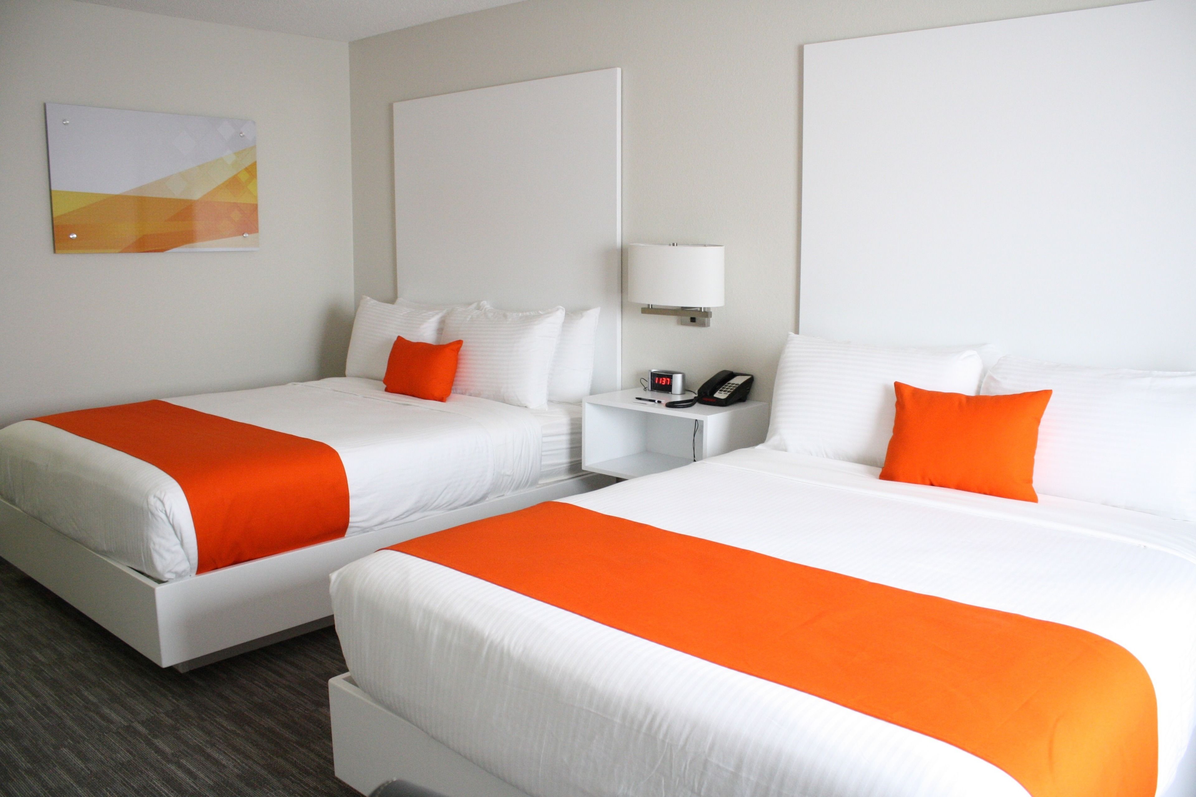A hotel room with two beds, white bedding, orange accents, a nightstand with a phone, and a wall-mounted lamp.