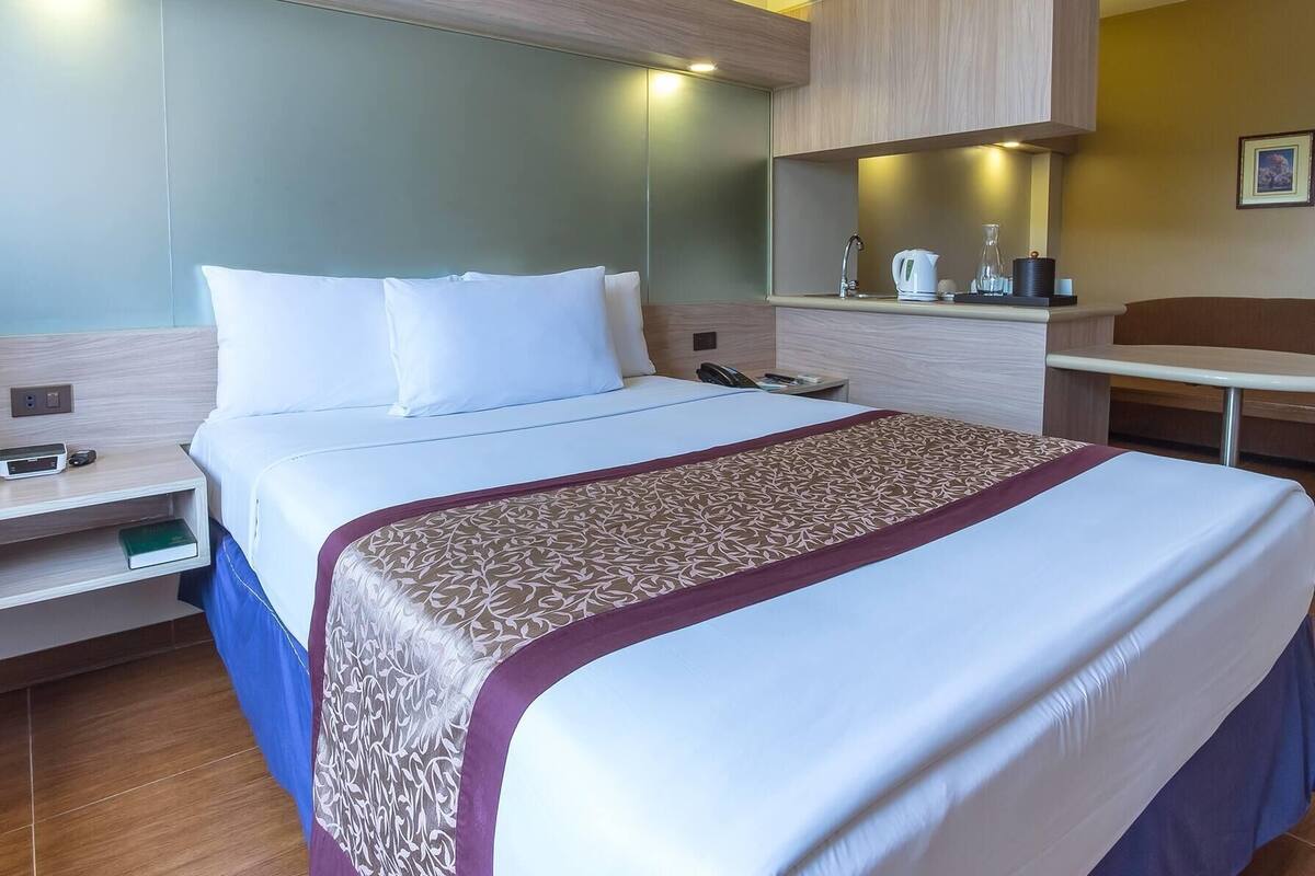 A hotel room with a large bed, a bedside table with a phone and a book, a desk with a chair, and a mirror above the desk.