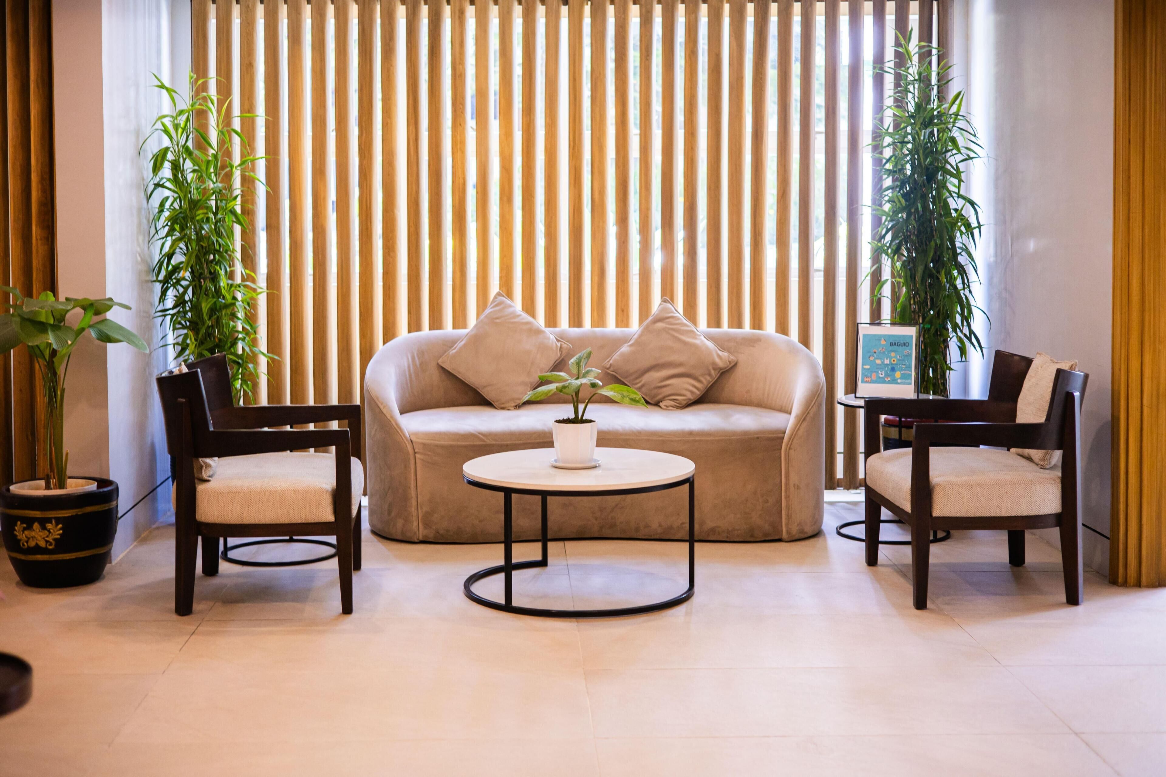 A room with a beige sofa, two wooden chairs, a small round table, and two potted plants.