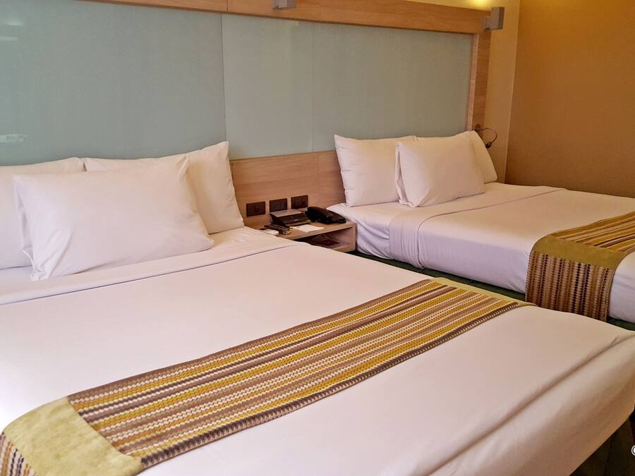 A hotel room with two beds, each with white linens and a striped blanket. There is a nightstand with a phone and a lamp between the beds.