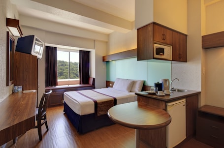 A hotel room with a bed, a desk, a small kitchenette, and a window with curtains.
