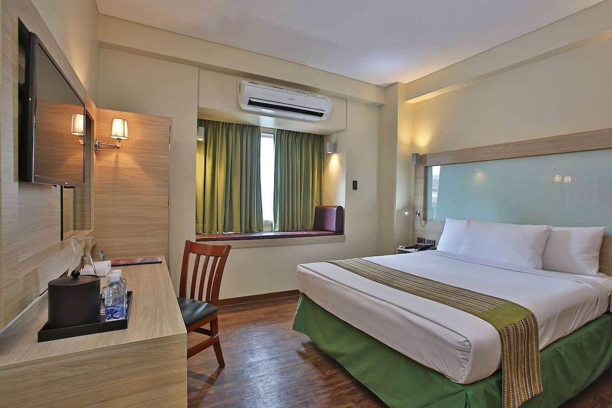A hotel room with a bed, a desk with a chair, a TV, and an air conditioning unit.