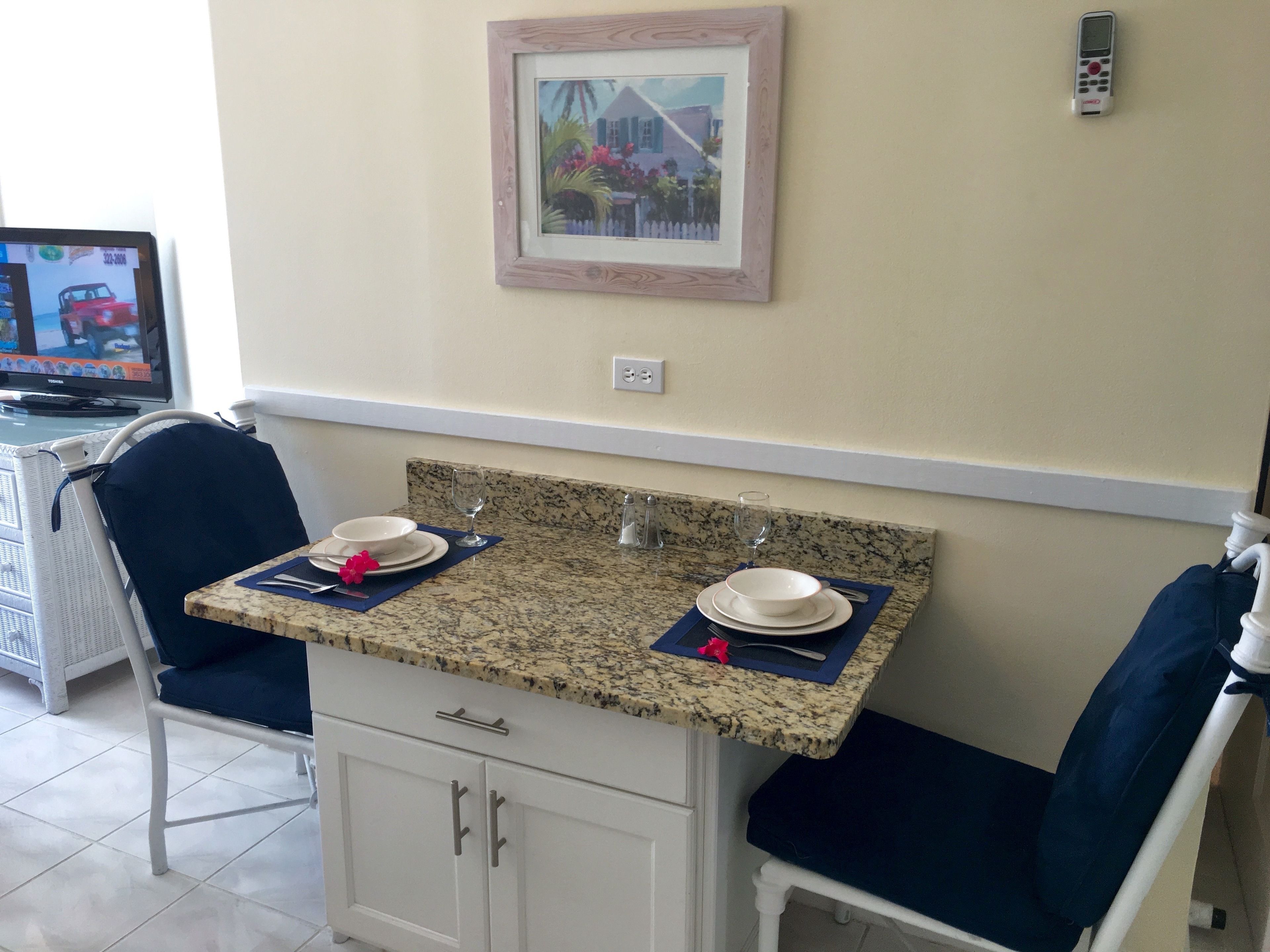 A dining area with a granite countertop, two white cabinets, a framed picture on the wall, and a television in the background.