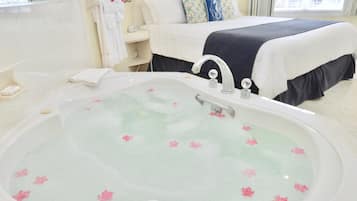 Une chambre d’hôtel avec une grande baignoire remplie d’eau et de pétales de rose, un lit avec du linge de lit blanc et des coussins décoratifs, et une salle de bain avec un miroir et des articles de toilette.