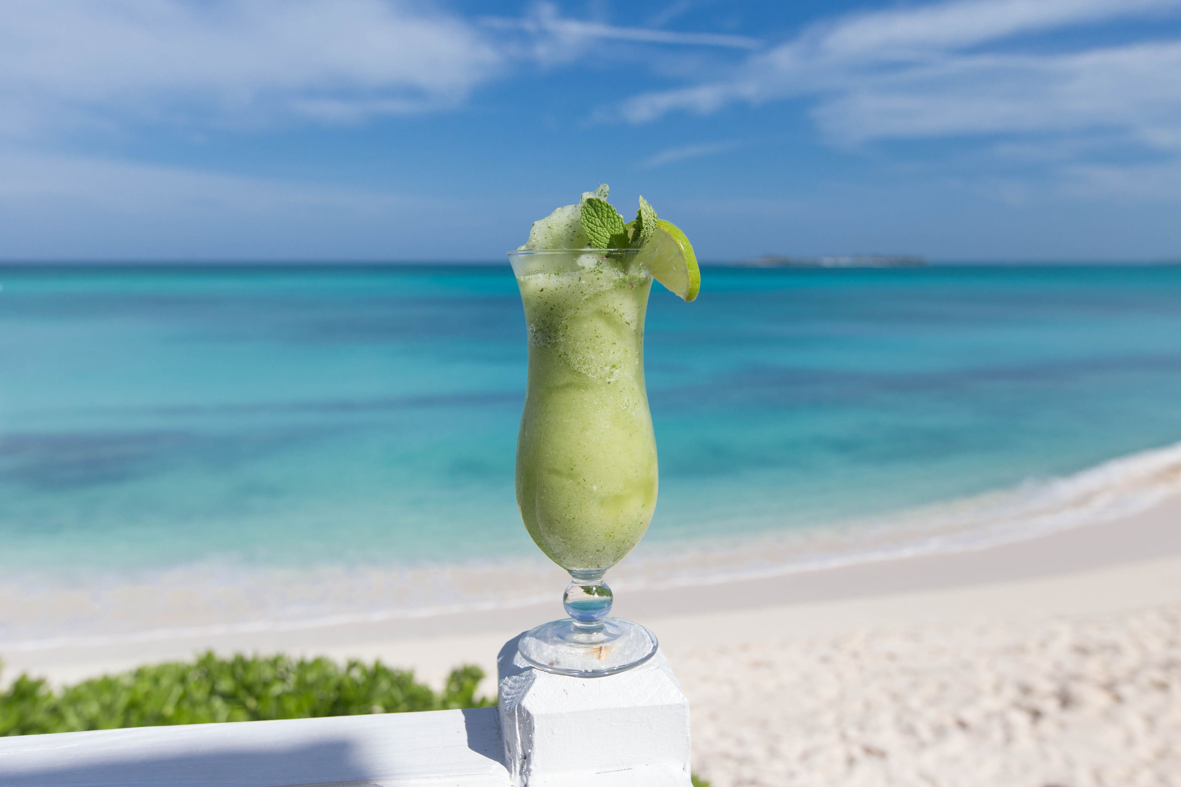 Un verre d’une boisson verte, agrémenté d’une tranche de citron vert, est posé sur une surface blanche, avec en arrière-plan une plage aux eaux turquoise et limpides et un rivage de sable blanc.