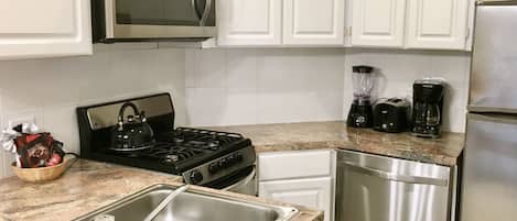 Una cocina con gabinetes blancos, microondas, estufa, fregadero, lavavajillas, licuadora y cafetera.
