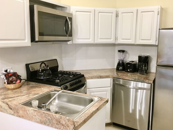Una cocina con gabinetes blancos, microondas, estufa, fregadero, lavavajillas, licuadora y cafetera.