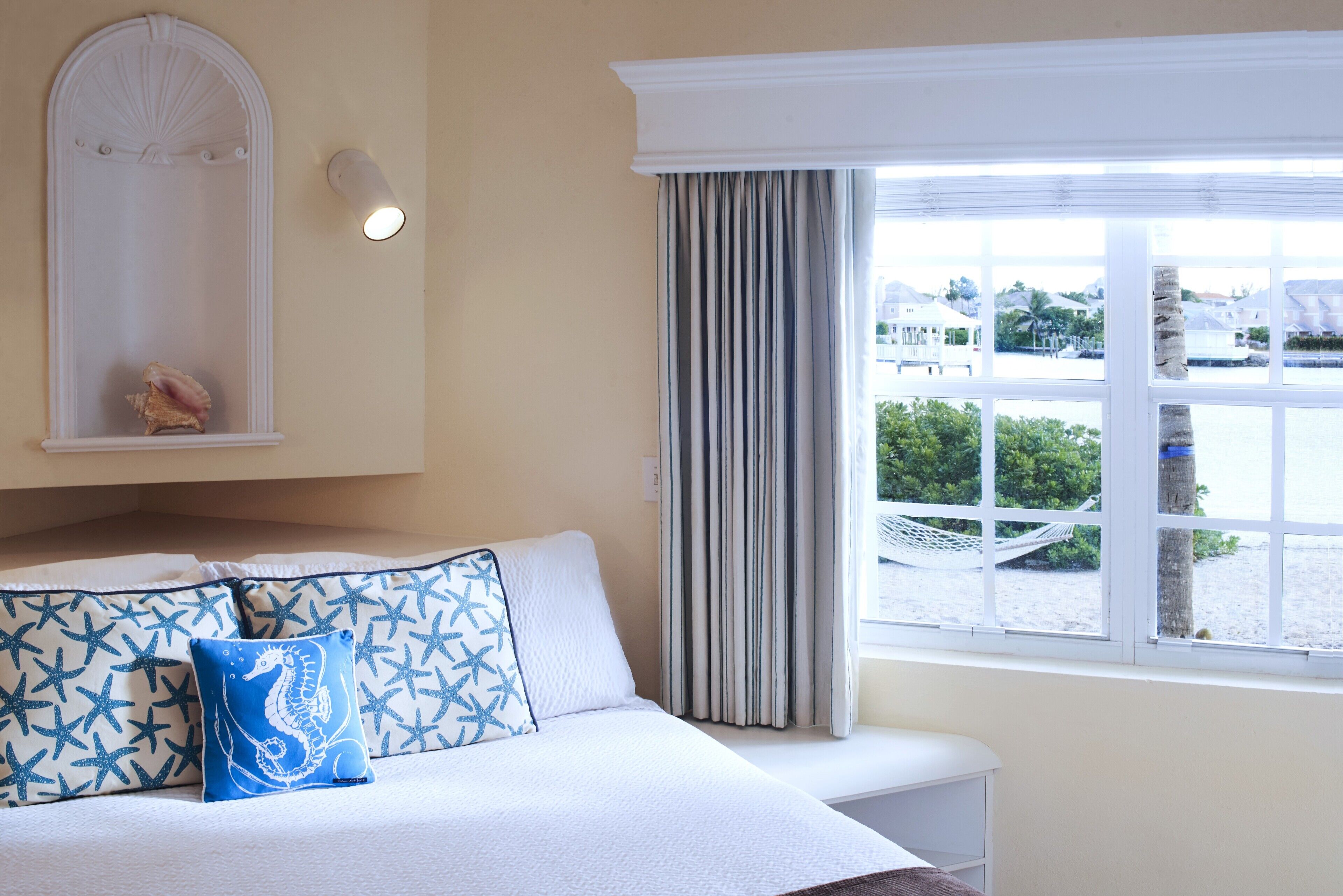 A bedroom with a bed, a window with curtains, a wall-mounted light, and a view of the beach.