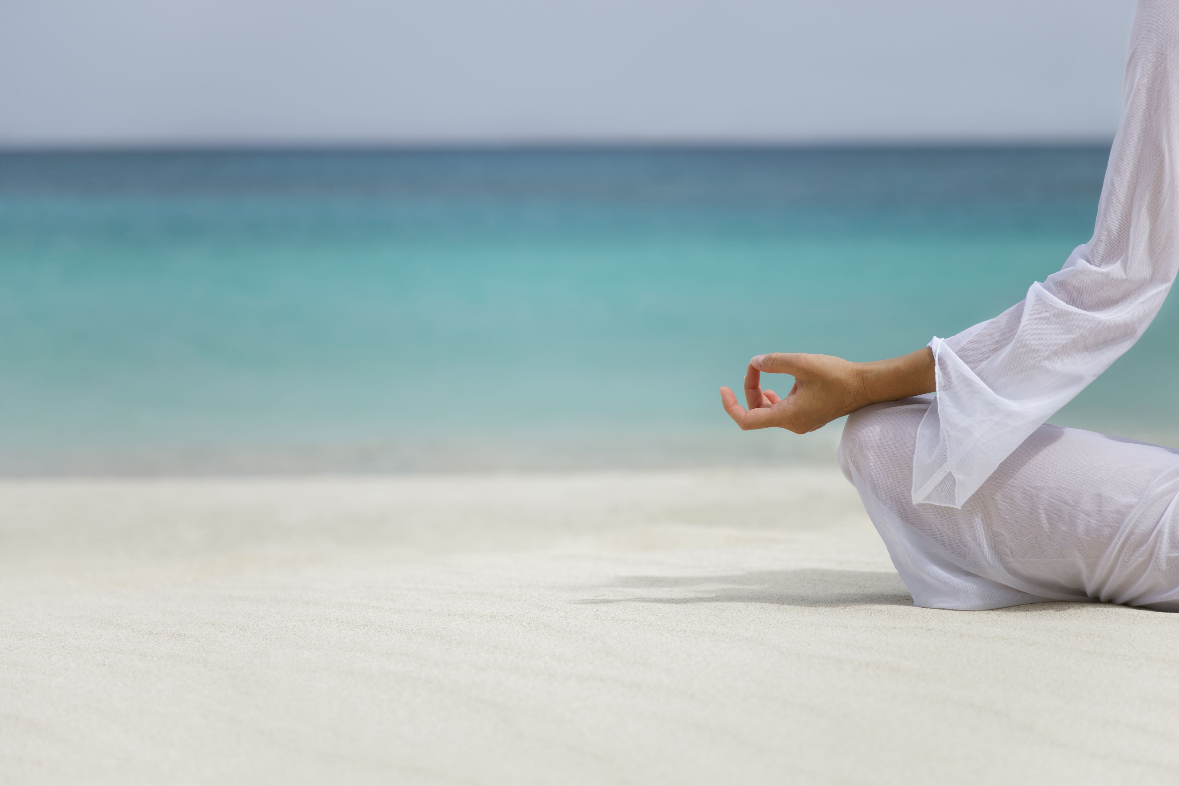 Une personne vêtue de blanc est assise sur une plage de sable, avec une mer calme en arrière-plan.