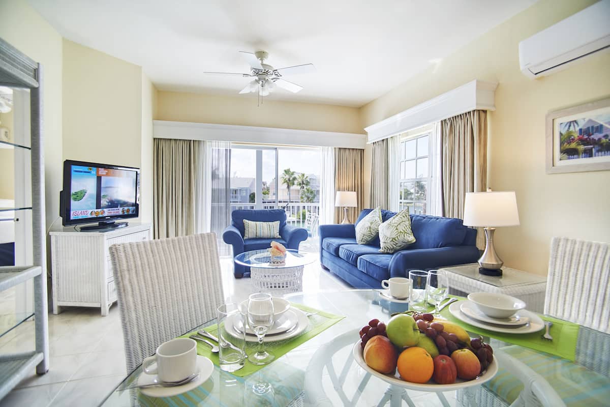 A hotel room with a living area featuring a blue sofa, a television, a dining table with fruit, and a balcony with curtains.