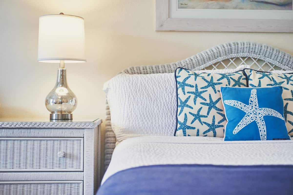 A neatly made bed with a white and blue starfish pillow, a white lamp on a wicker nightstand, and a framed picture on the wall.