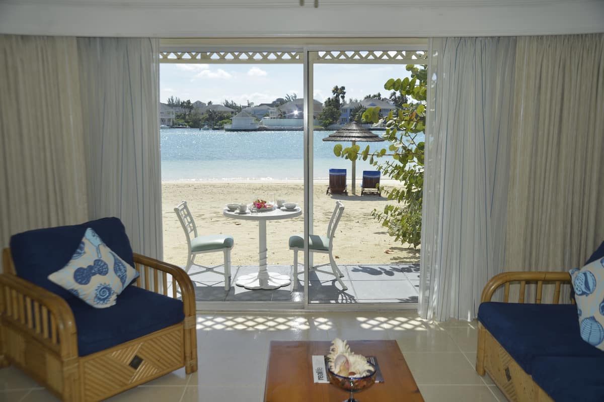 A room with a view of a beach, two wicker sofas with cushions, a small table with a bowl and a glass, and a patio area with chairs and a table.