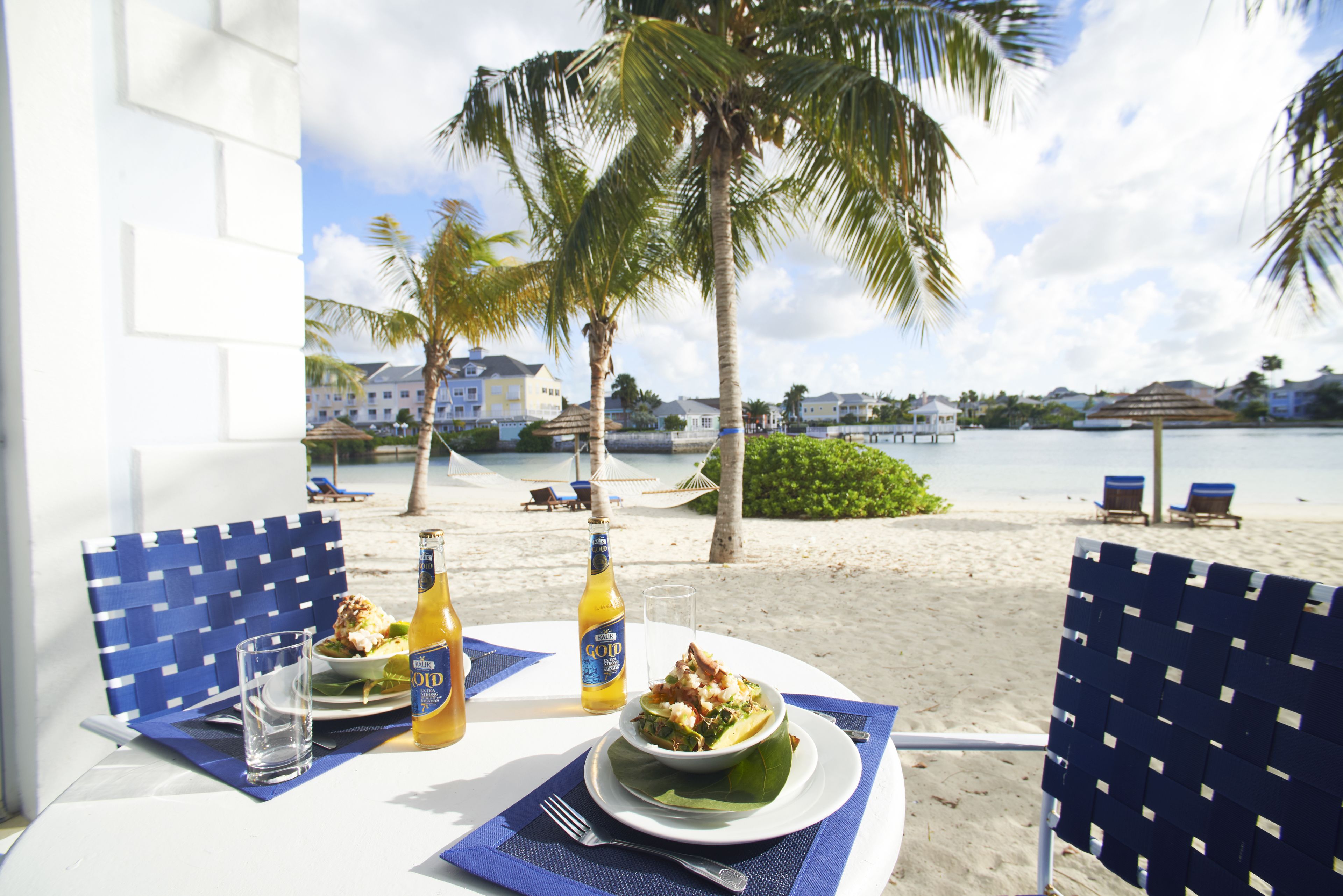 A dining table with a meal, two beer bottles, and a glass of water on a balcony overlooking a beach with palm trees and lounge chairs.