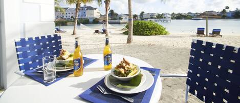 A dining table with a meal, two beer bottles, and a glass of water on a balcony overlooking a beach with palm trees and lounge chairs.