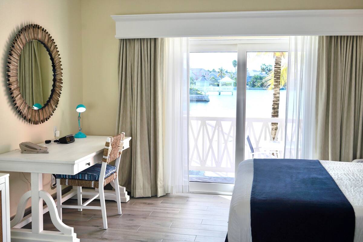 A hotel room with a bed, a desk with a chair, a phone, a lamp, and a view of the outside.