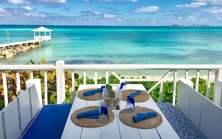 A table with blue cushions and woven placemats, overlooking a turquoise sea and a white gazebo on a pier.