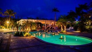 A swimming pool with a covered seating area, palm trees, and a building with a covered patio in the background. - Beach House - Adults Only (Providenciales)