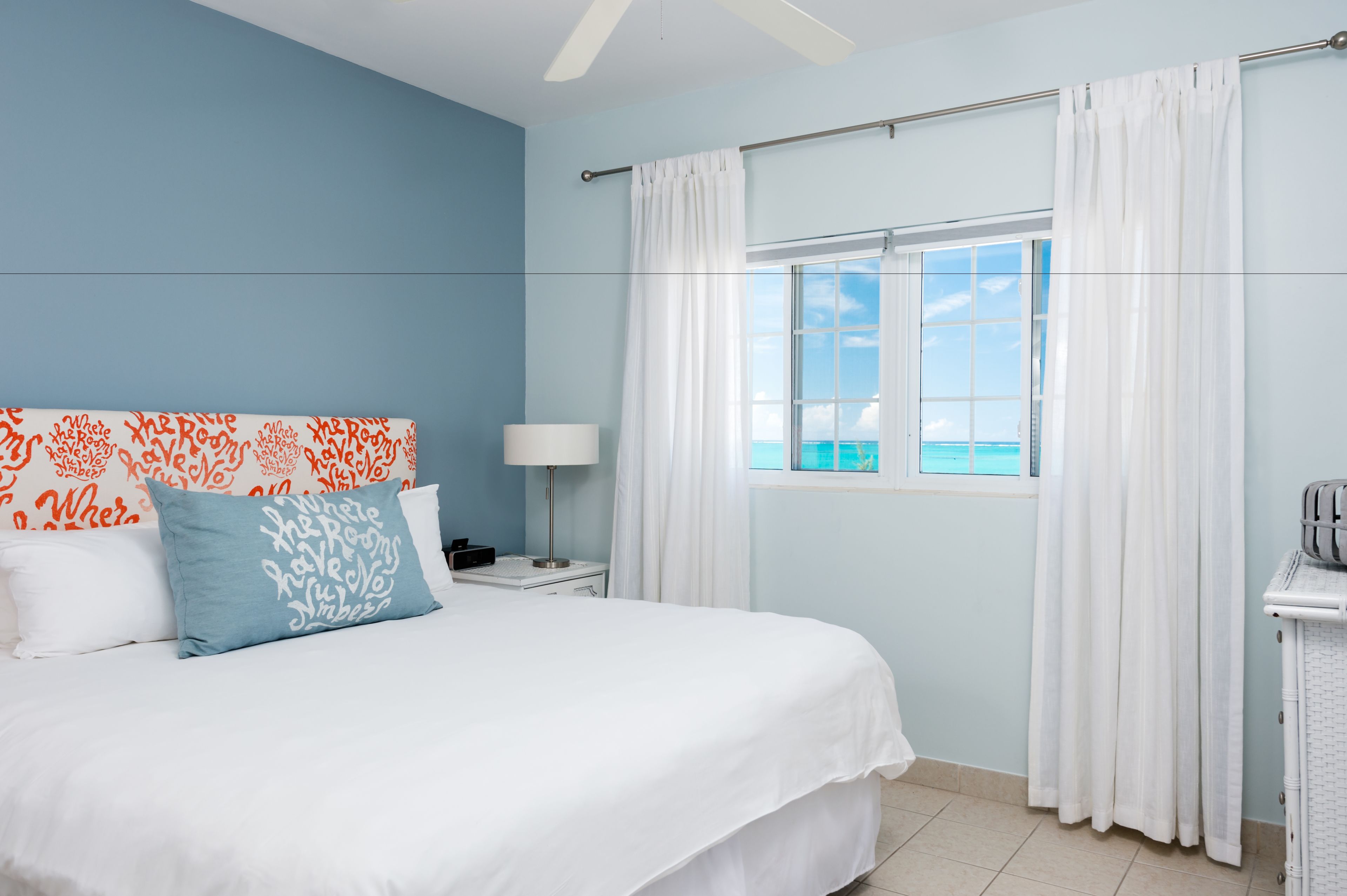 A bedroom with a bed, a nightstand, a lamp, a window with curtains, and a view of the ocean.