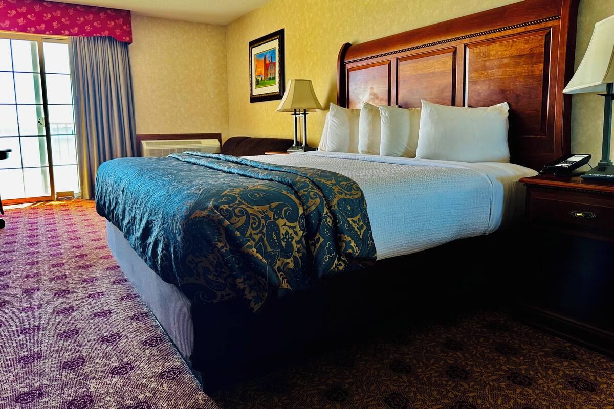 A hotel room with a large bed, a wooden headboard, a nightstand with a lamp, and a window with curtains.