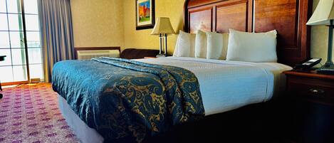 A hotel room with a large bed, a wooden headboard, a nightstand with a lamp, and a window with curtains.