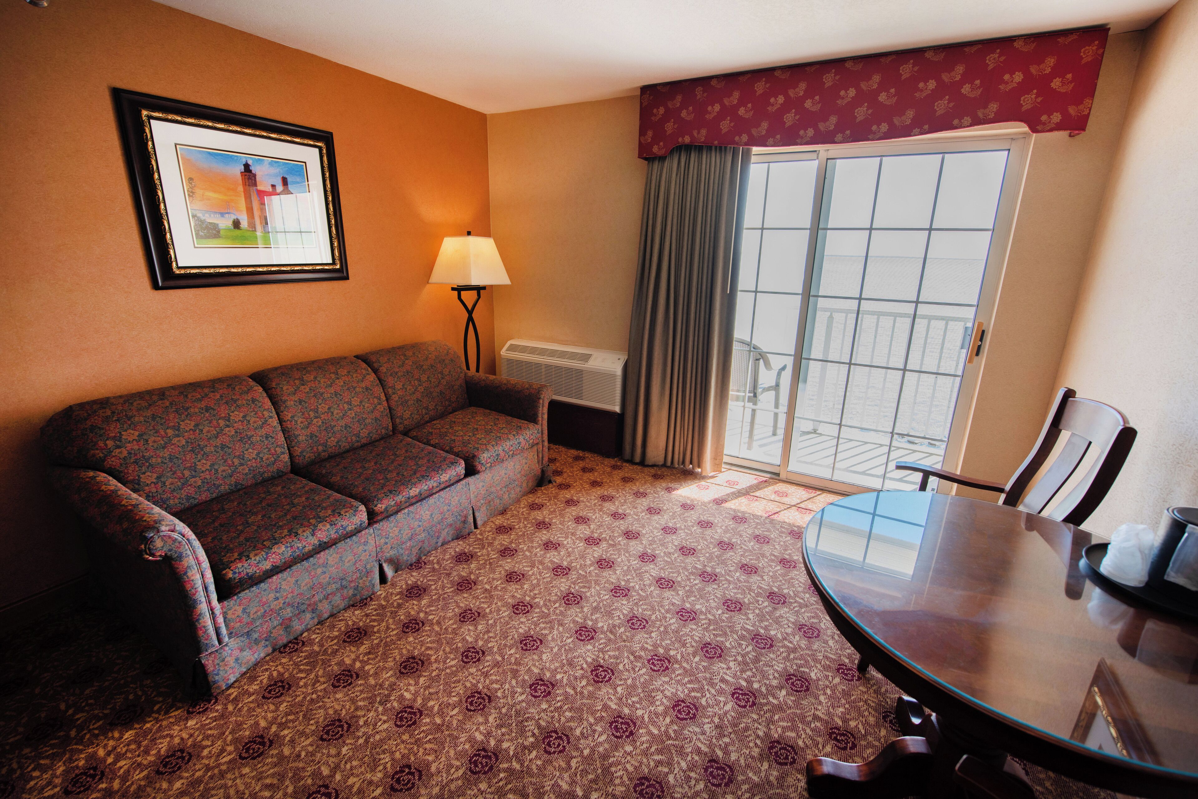 A hotel room with a patterned carpet, a sofa, a table with chairs, a lamp, a framed picture on the wall, and a window with curtains.