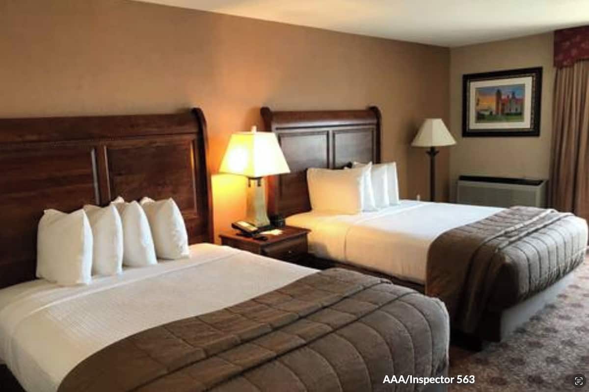 A hotel room with two beds, wooden headboards, bedside lamps, a nightstand, and a framed picture on the wall.
