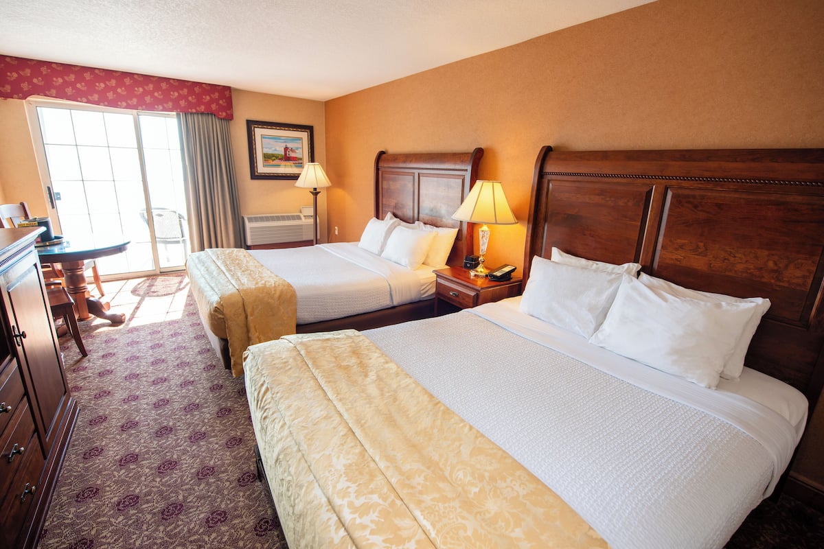 A hotel room with two beds, a desk, and a chair. There is a window with curtains, a painting on the wall, and a lamp on the nightstand.