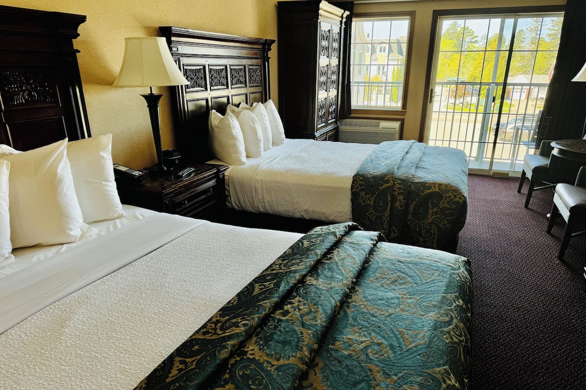 A hotel room with two beds, a nightstand, a lamp, and a view of the outside through a window.