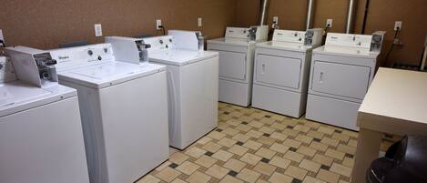 A laundry room with multiple white washers and dryers, a tiled floor, and a trash can.