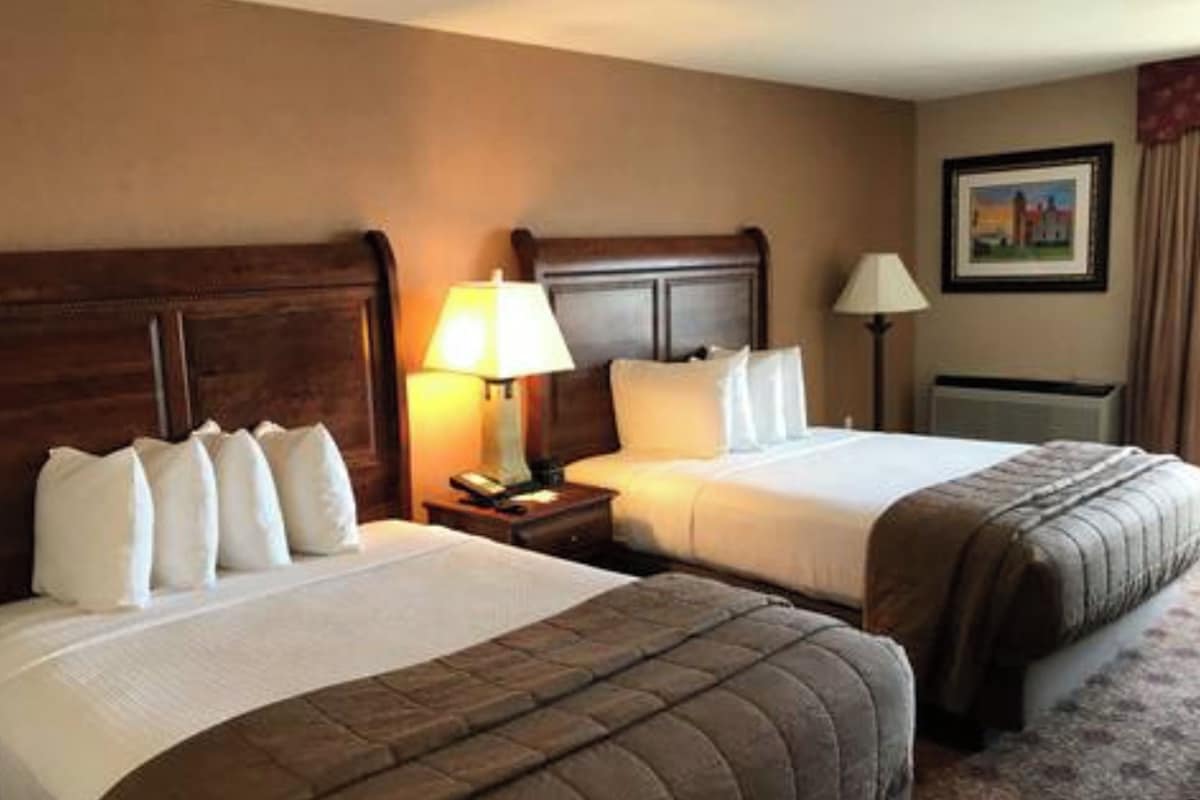 A hotel room with two beds, wooden headboards, bedside lamps, a nightstand, and a framed picture on the wall.