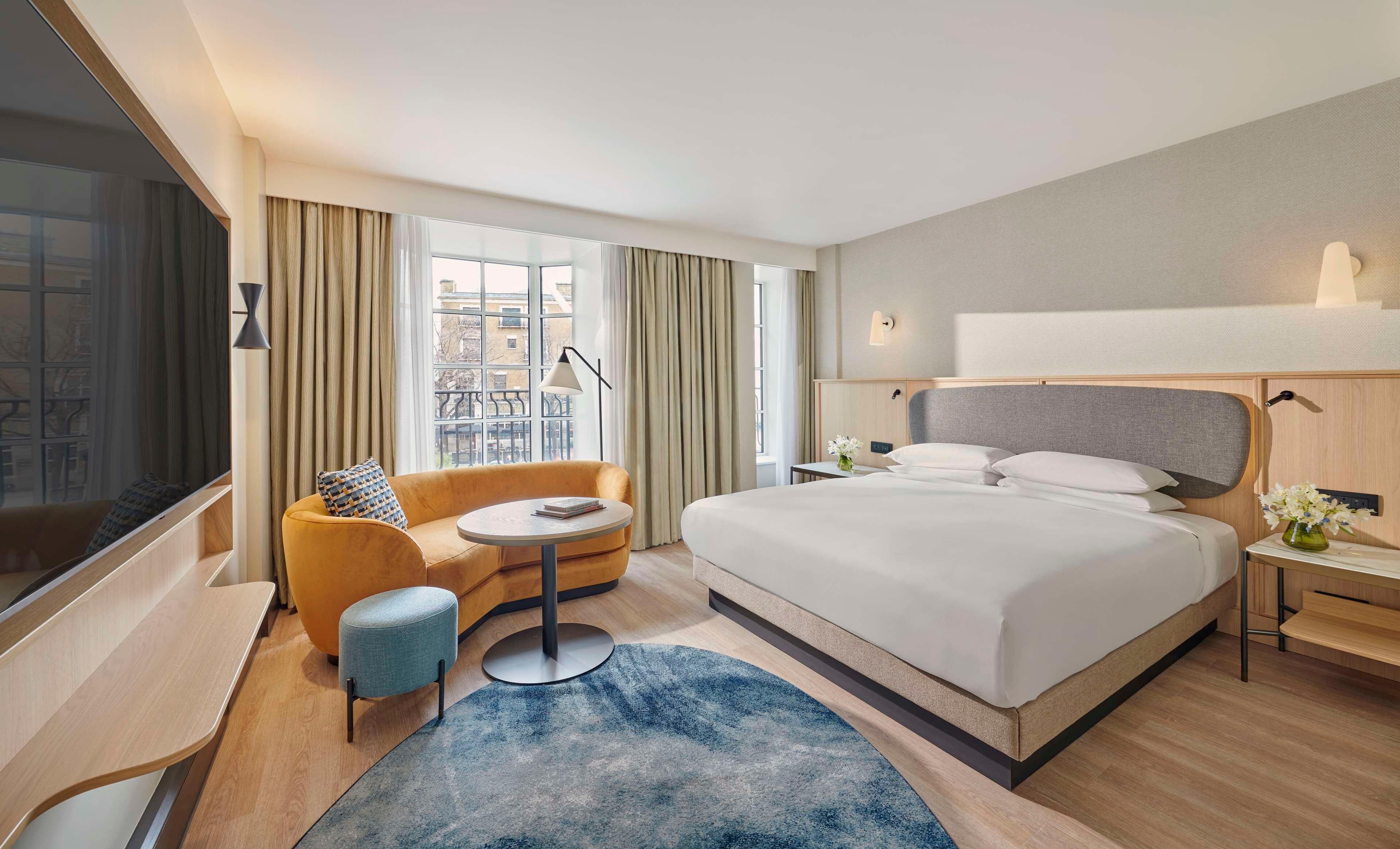 Une chambre d’hôtel avec un grand lit, un canapé, une petite table ronde et une vue sur la ville grâce à de grandes fenêtres.