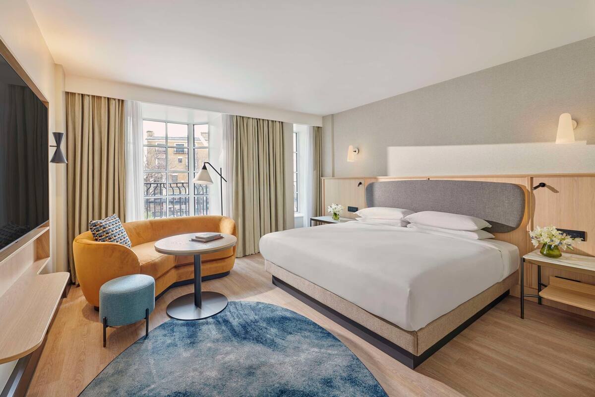 Une chambre d’hôtel avec un grand lit, un canapé, une petite table ronde et une vue sur la ville grâce à de grandes fenêtres.