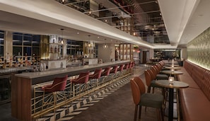 Un bar moderno con una barra larga, taburetes y una zona de estar con mesas y sillas.