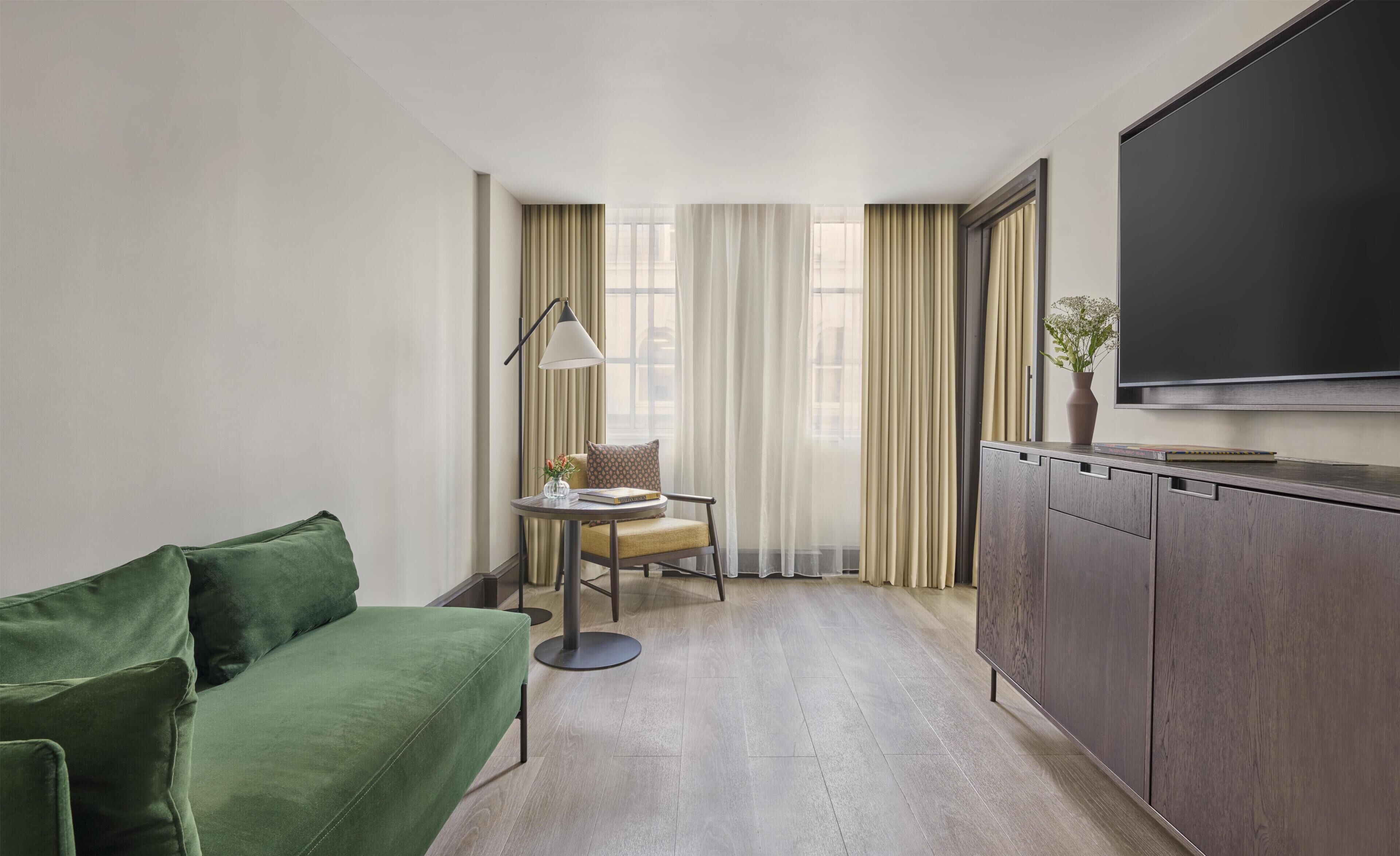 Una habitación de hotel moderna con un sofá verde, una mesita con una lámpara, una silla, un televisor y una ventana con cortinas.