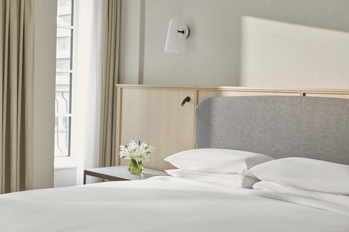 Une chambre d’hôtel avec un lit, une table de chevet ornée d’un vase de fleurs, une lampe fixée au mur et une fenêtre avec des rideaux.