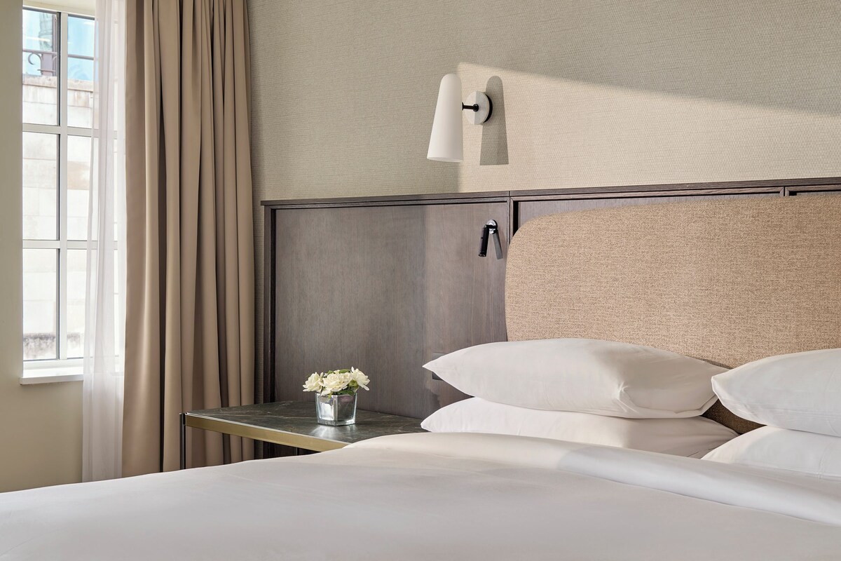 Una habitación de hotel con cama, mesita de noche, lámpara de pared, ventana con cortinas y un pequeño florero con flores.