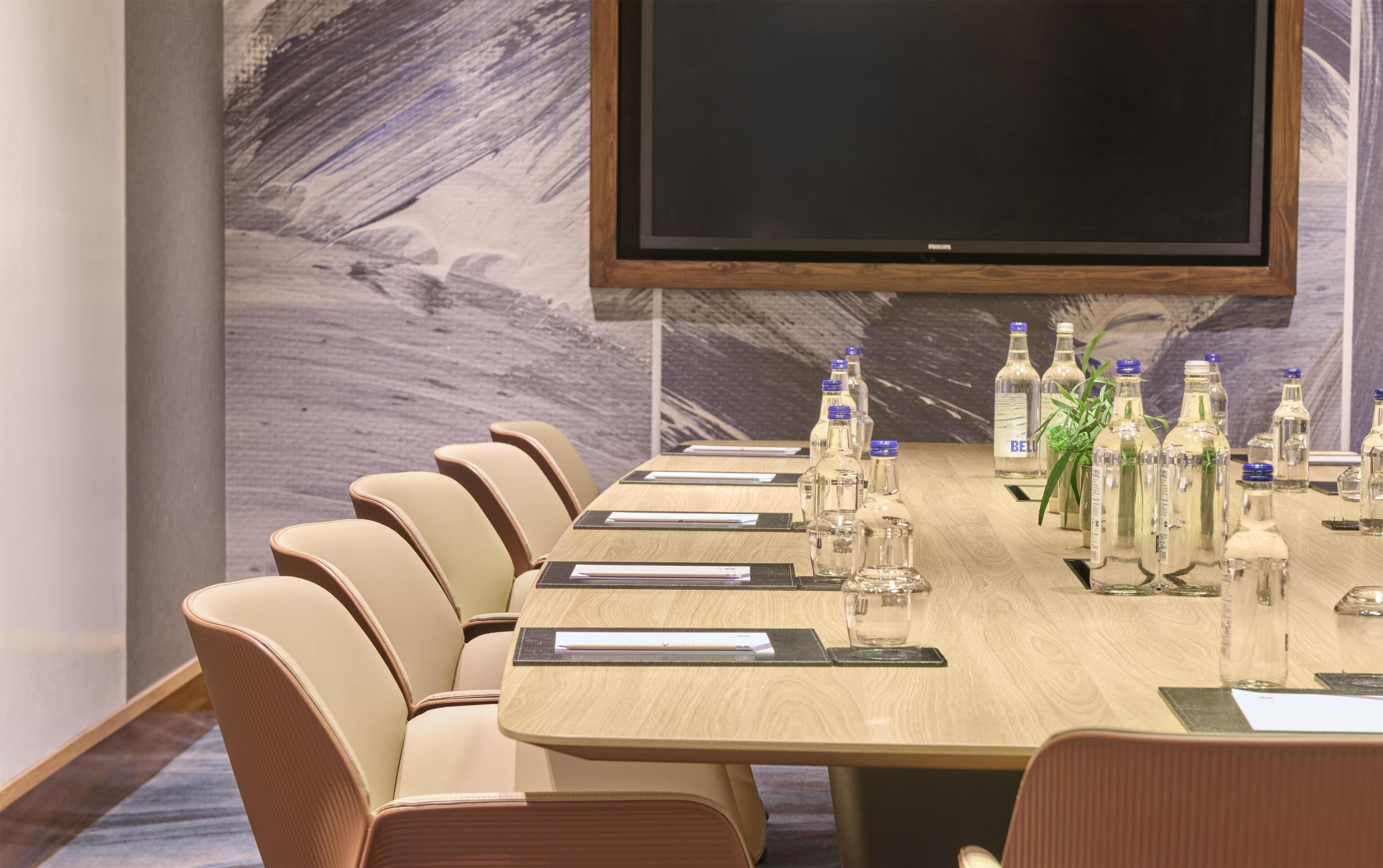 A conference room with a long table, beige chairs, and a large flat-screen TV on the wall.