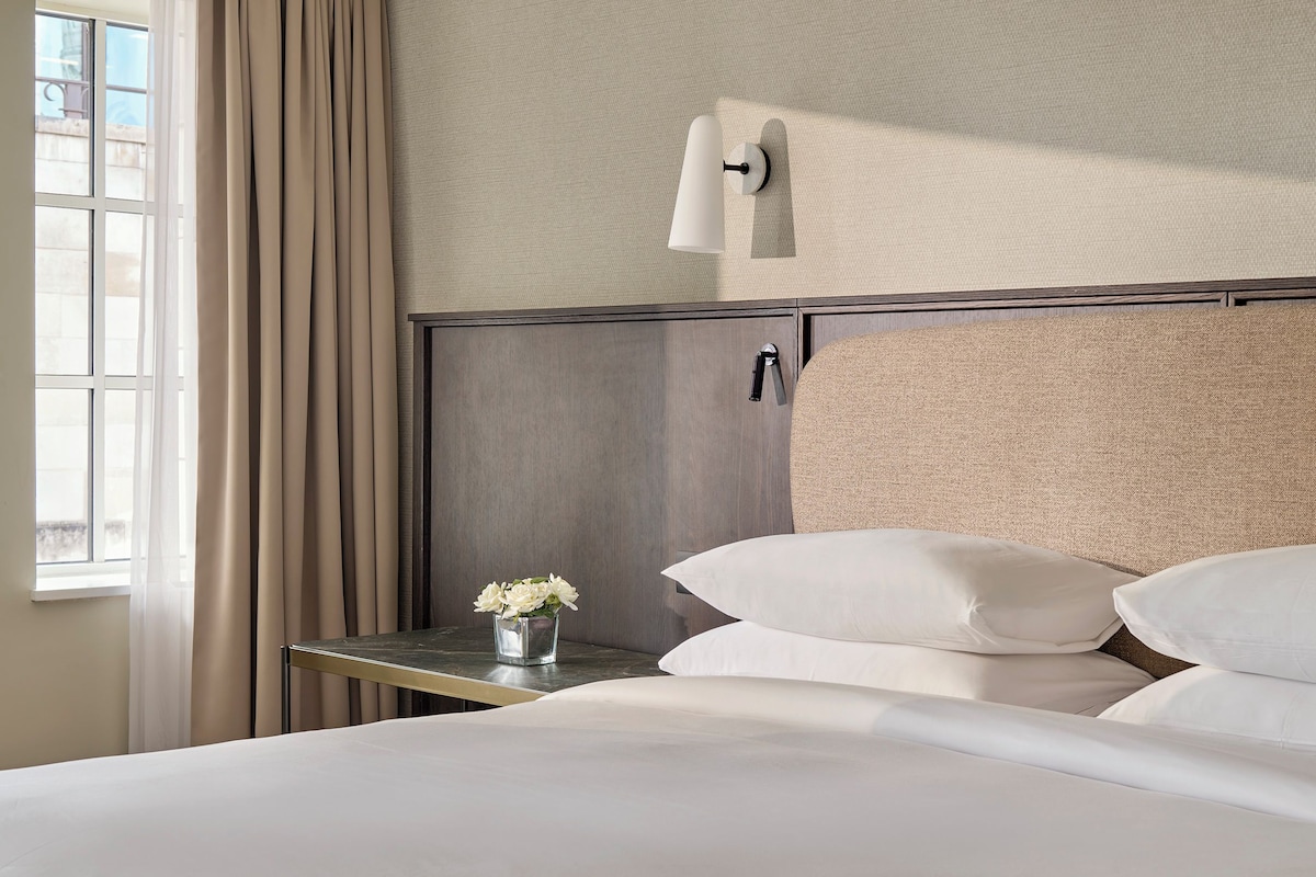 A hotel room with a bed, a nightstand, a wall-mounted lamp, a window with curtains, and a small vase with flowers.