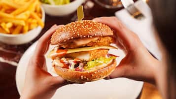 A person holding a hamburger with a sesame seed bun, surrounded by a dining table with a white plate, a bowl of fries, and a glass of wine.