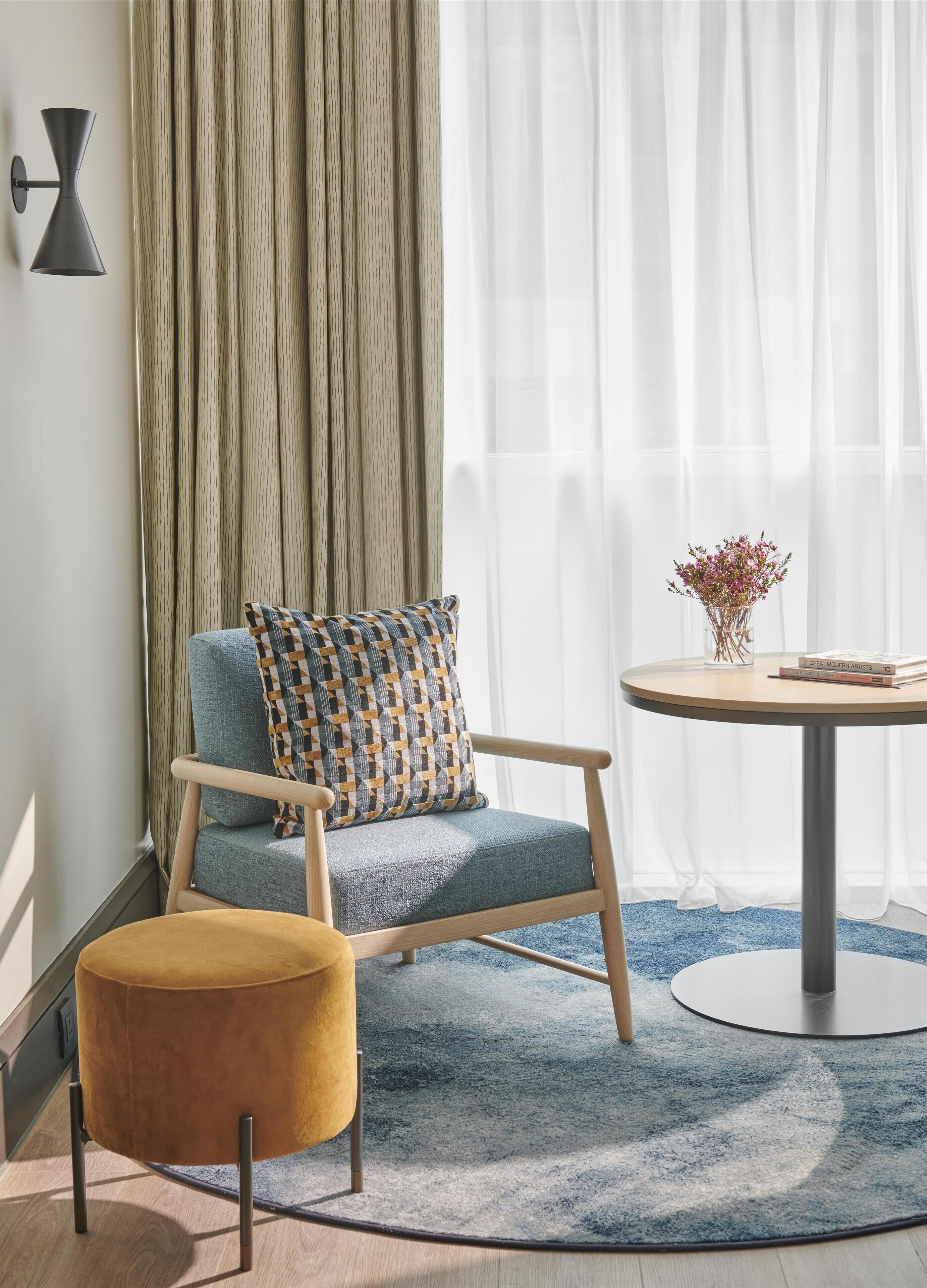 Een moderne kamer met een blauwe bank, een klein rond tafeltje met een bloevas, en een groot raam met dunne gordijnen.