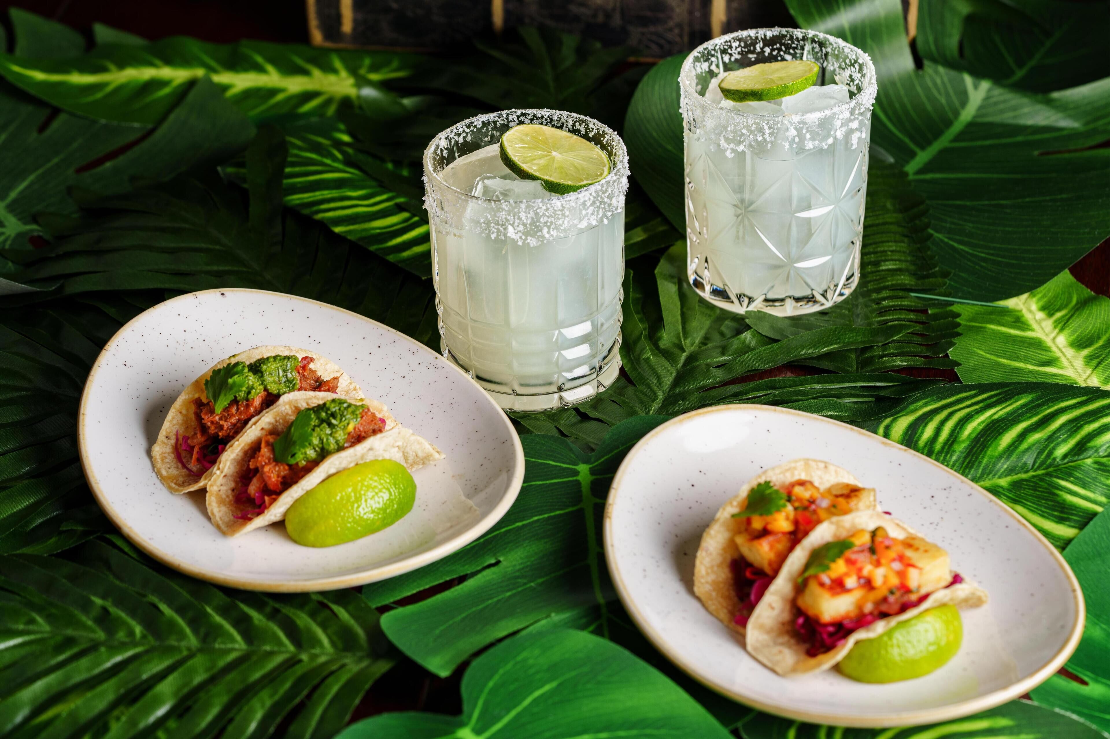Two plates of tacos and two glasses of a lime-based drink on a table with green leaves.