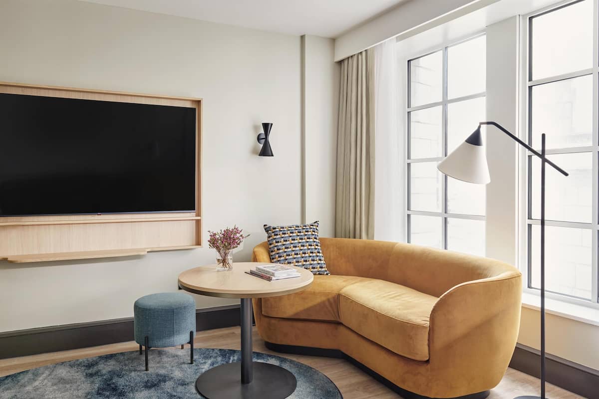 Un salon moderne avec un grand téléviseur à écran plat, une table basse ronde, un canapé de couleur moutarde, un petit pouf bleu et une lampe sur pied.