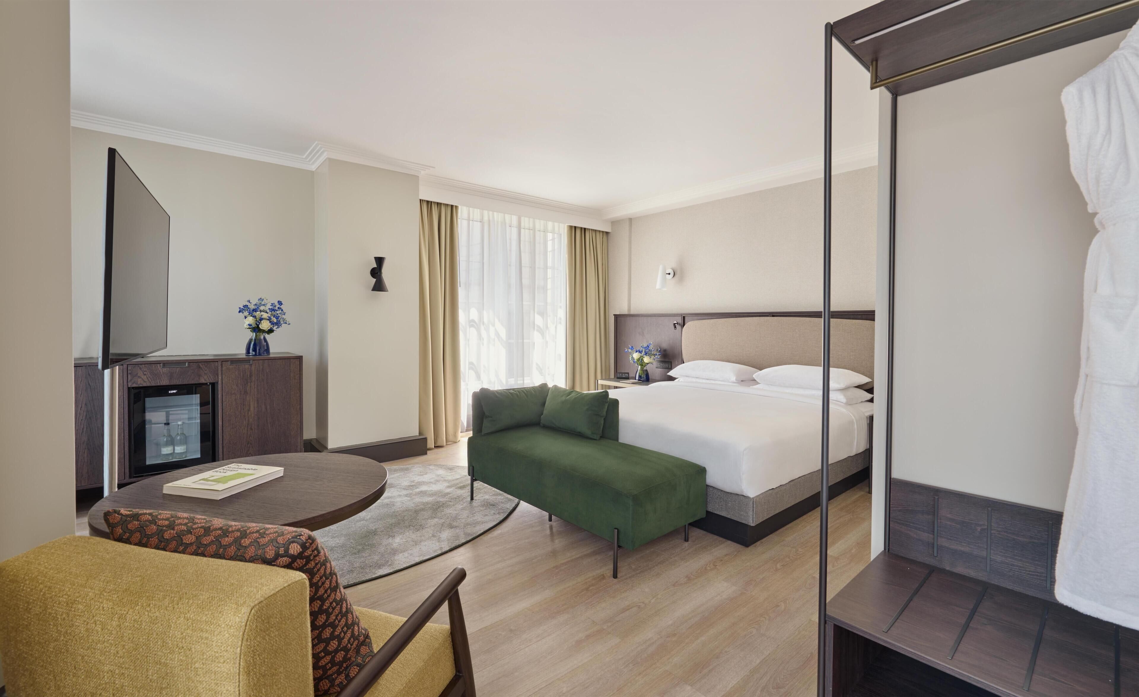Una habitación de hotel con una cama, un sofá verde, una mesita con un libro, un minibar y un espejo grande.
