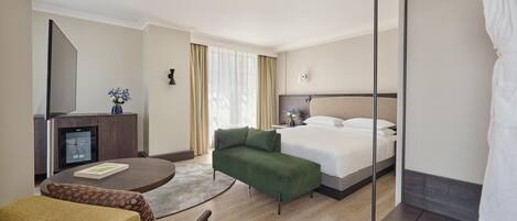 Una habitación de hotel con una cama, un sofá verde, una mesita con un libro, un minibar y un espejo grande.