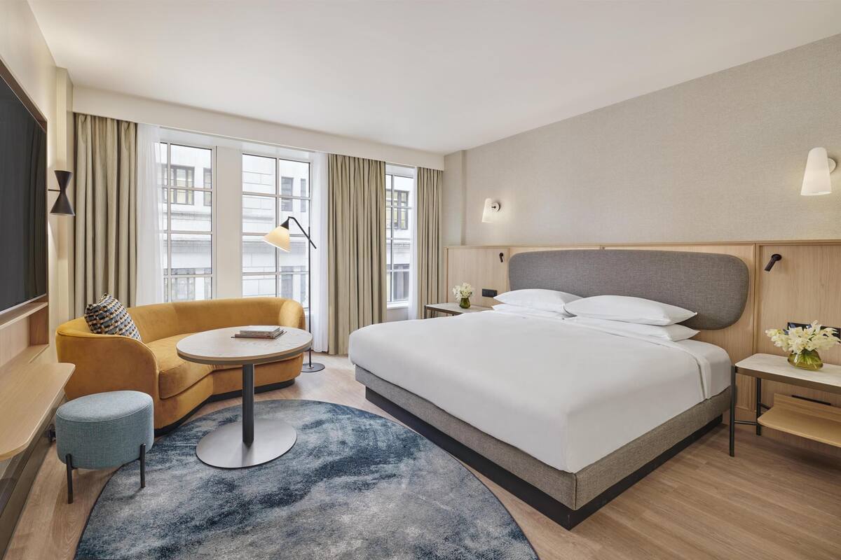 Une chambre d’hôtel avec un lit, un canapé, une petite table, une télévision et une fenêtre avec des rideaux.