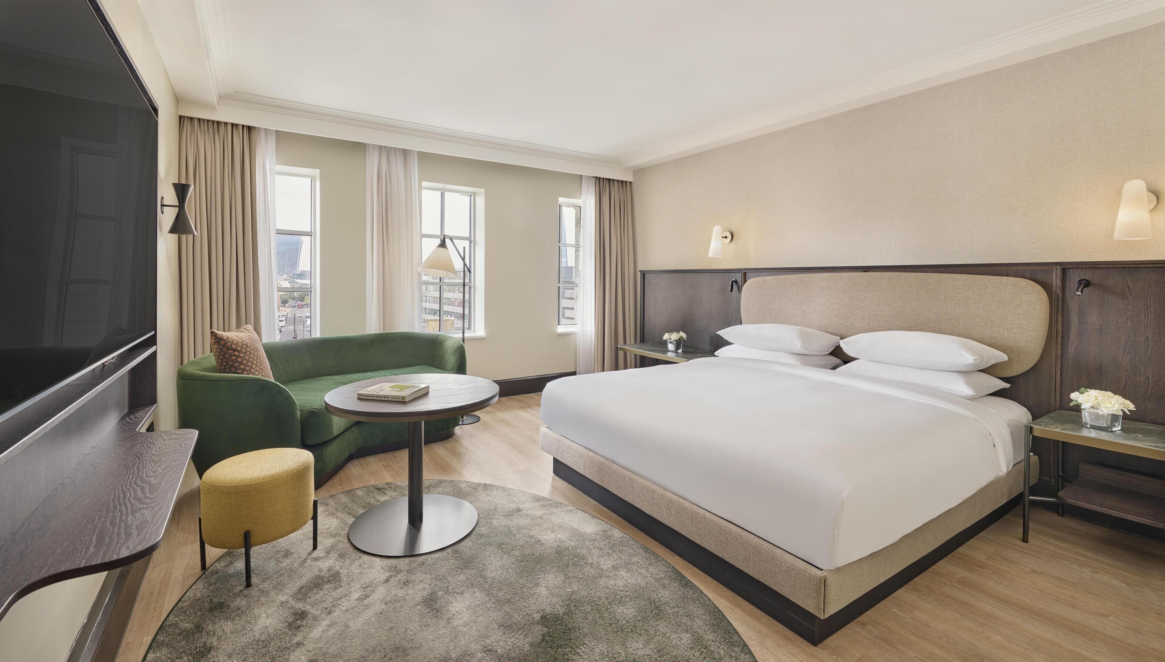 A hotel room with a large bed, a green sofa, a small round table, a TV, and a nightstand with a lamp.